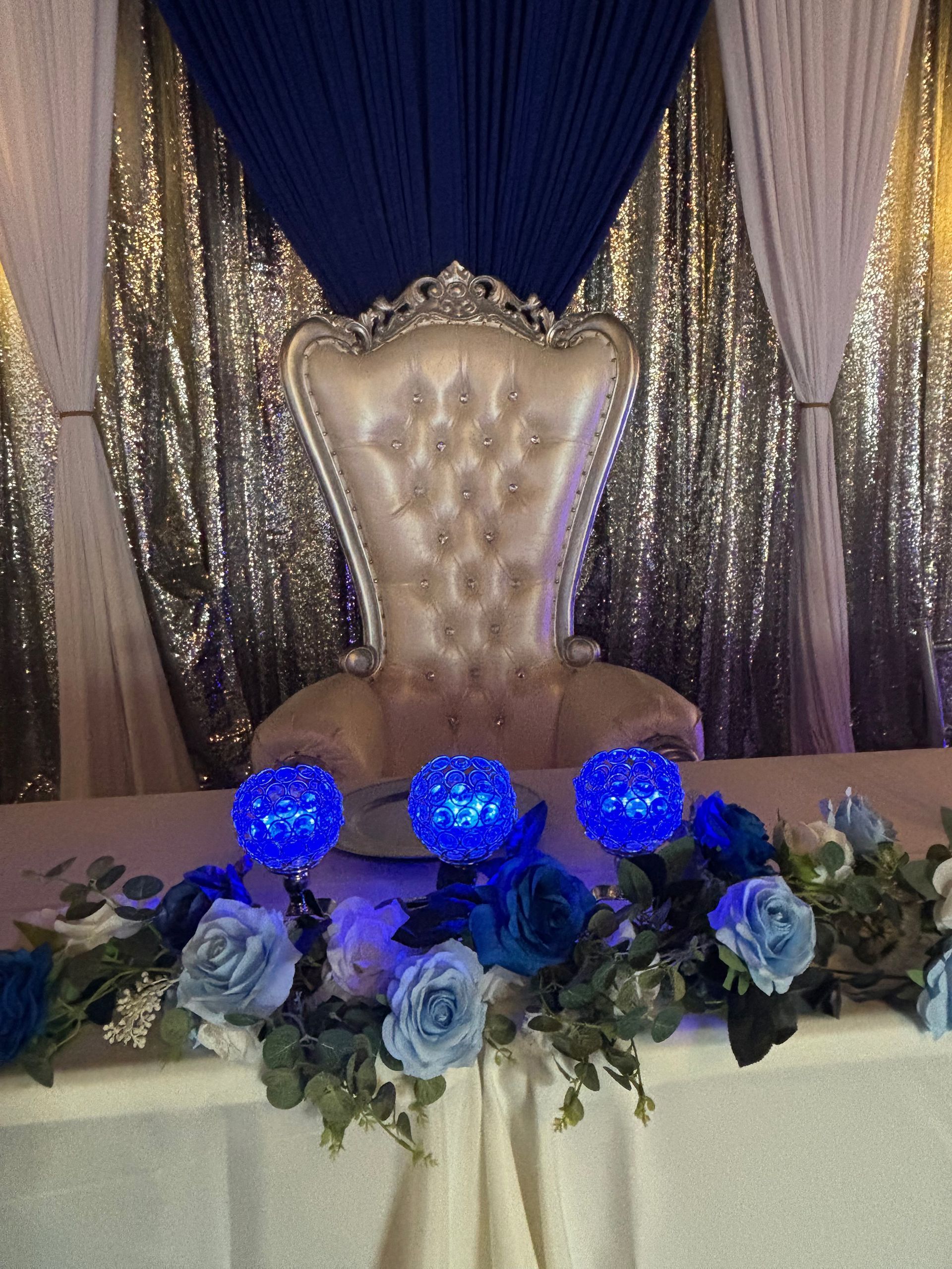 Gold throne chair with blue drapes, silver backdrop, and blue rose floral arrangement with glowing blue lights