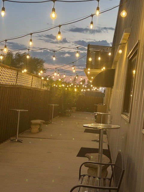 String-lit outdoor patio at dusk with tables, chairs, and a wooden fence beside a building
