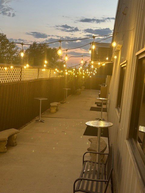 String lights illuminate a narrow outdoor patio at dusk beside a building, with tables and chairs along the wall.