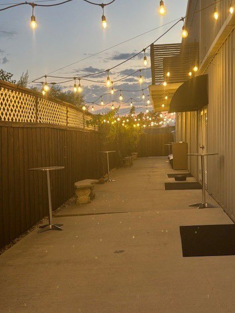String lights over a narrow outdoor alley at dusk, between a wooden fence and a building.