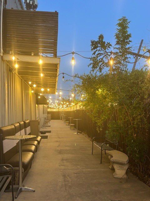 String lights illuminate a narrow outdoor patio with benches, tables, and plants at dusk.