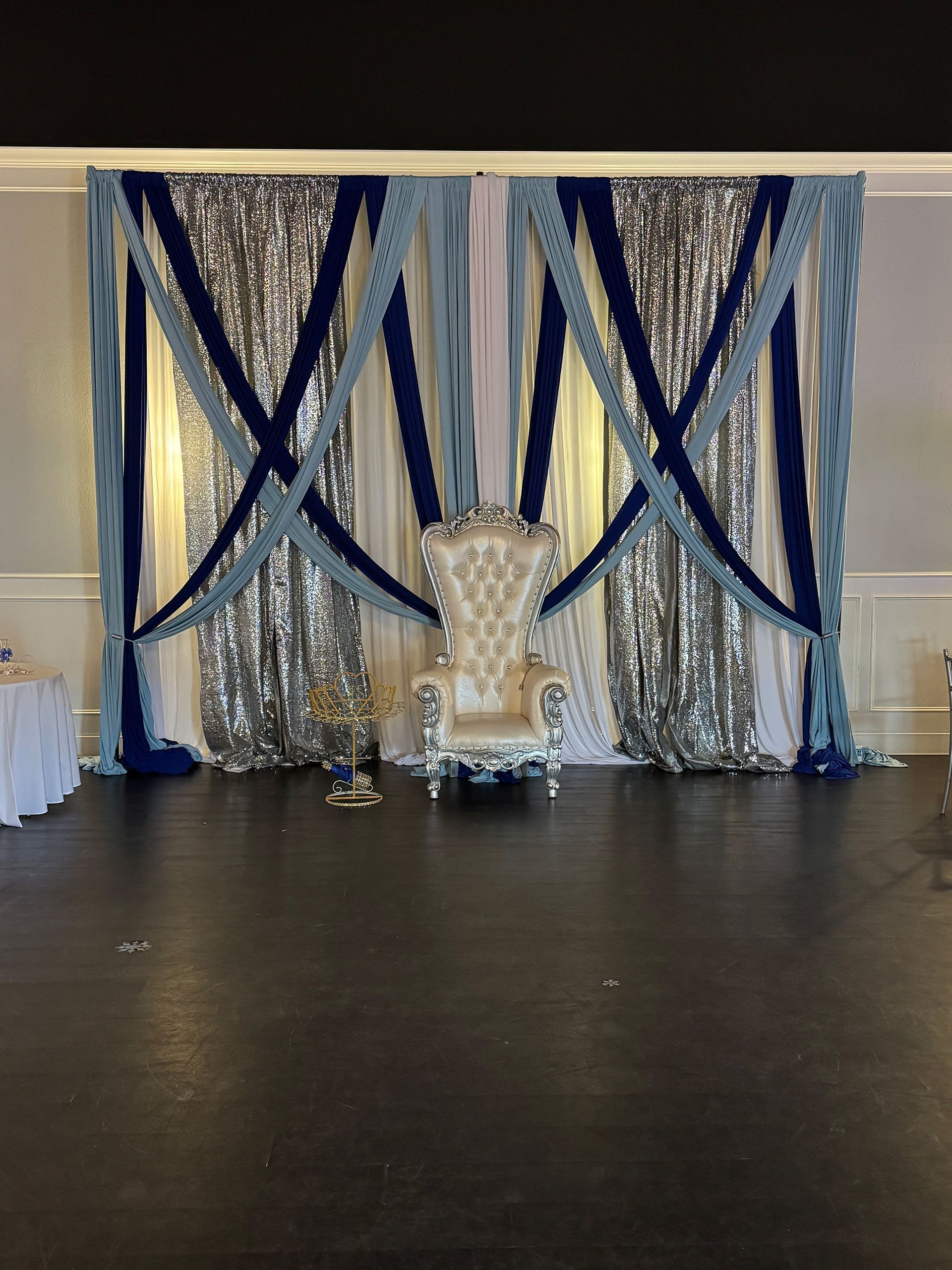 Blue and silver draped event backdrop with a white chair centerpiece on a dark floor