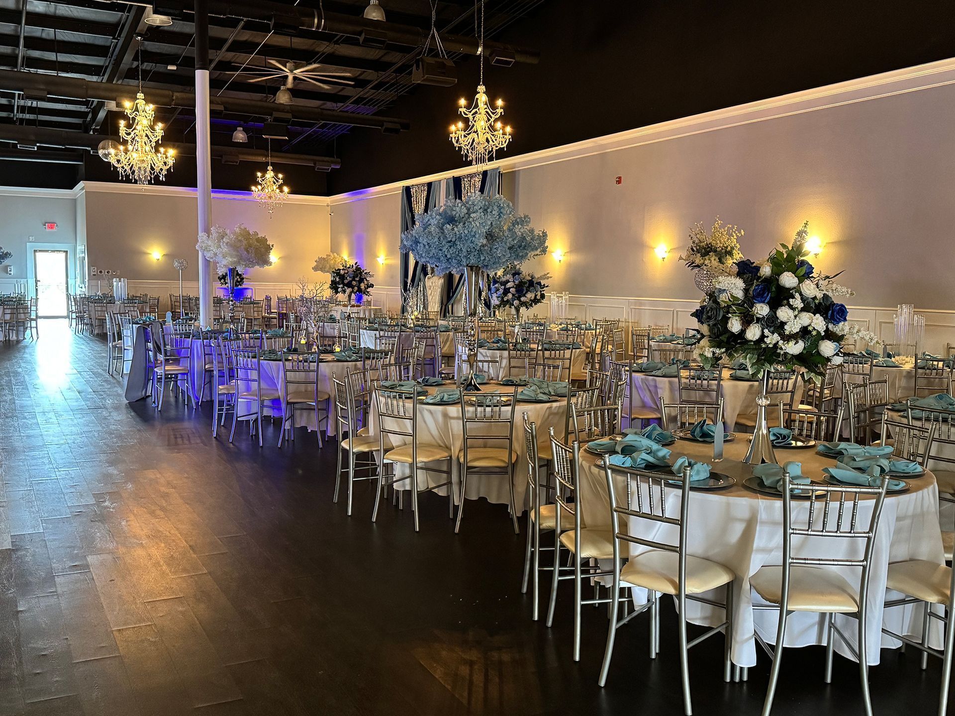 Elegant banquet hall with round tables, white chairs, and blue floral centerpieces under chandeliers