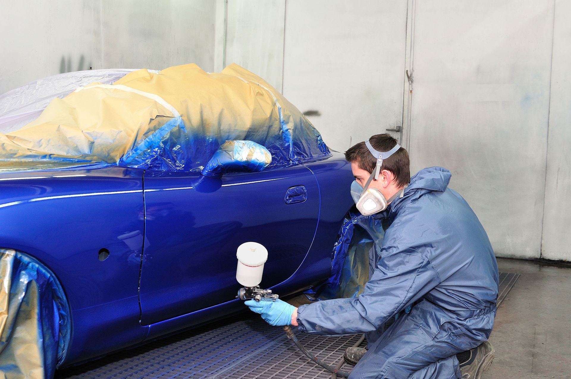 Person in protective suit spray-painting blue car in a paint booth.