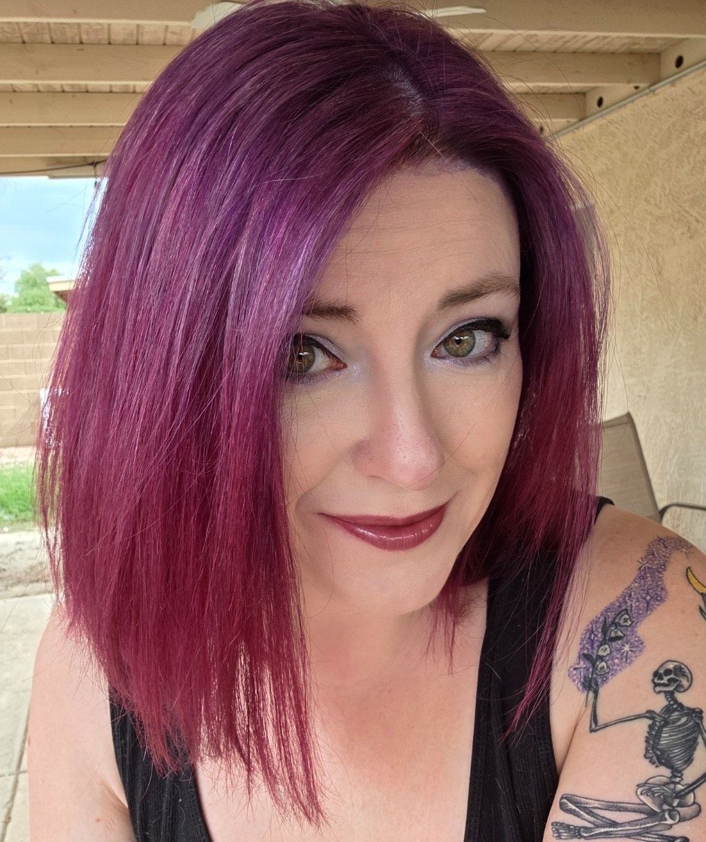 A woman with purple hair and dark lipstick, smiling. She has a shoulder tattoo in an outside setting.