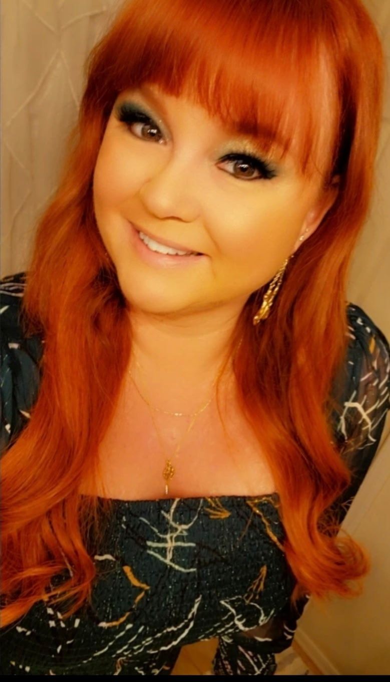 A woman with red hair, smiling, wearing a dark top, and eye makeup.