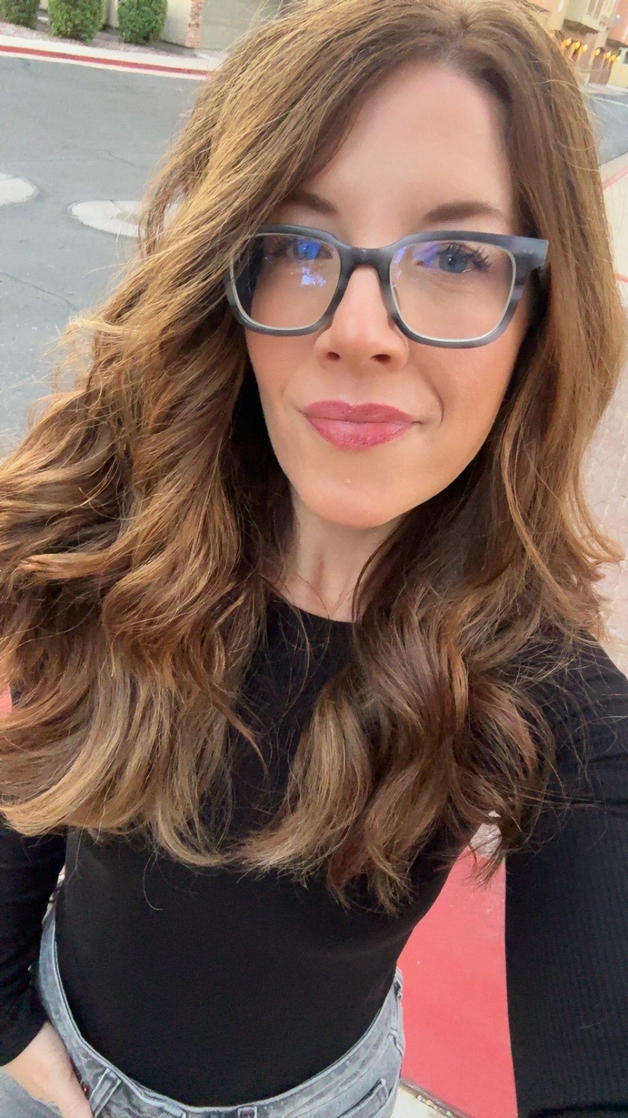 A woman with brown hair and glasses smiles. Black shirt. Outdoor setting.