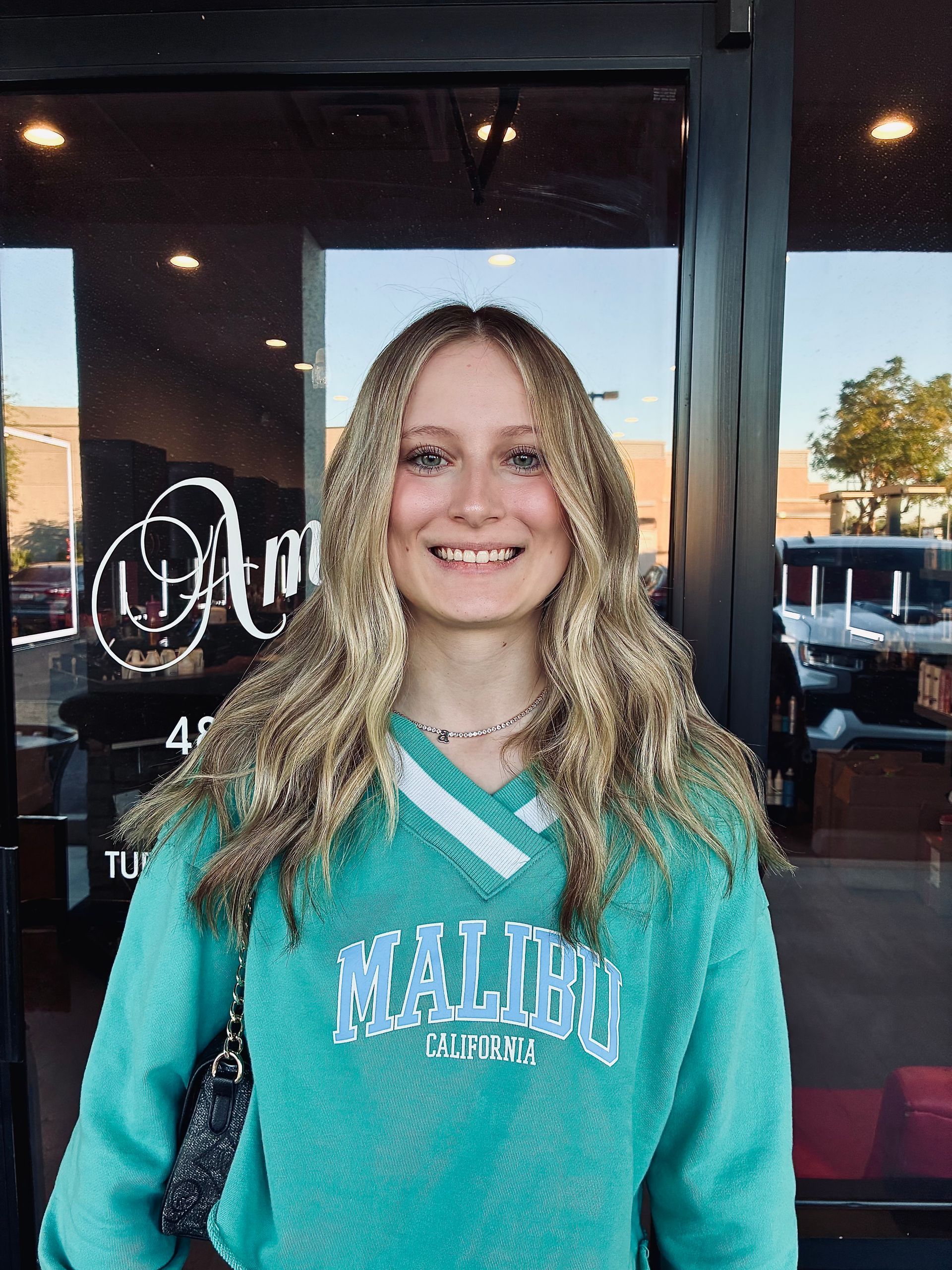 Woman with long blonde hair, smiling in front of a glass door, wearing a turquoise sweater with