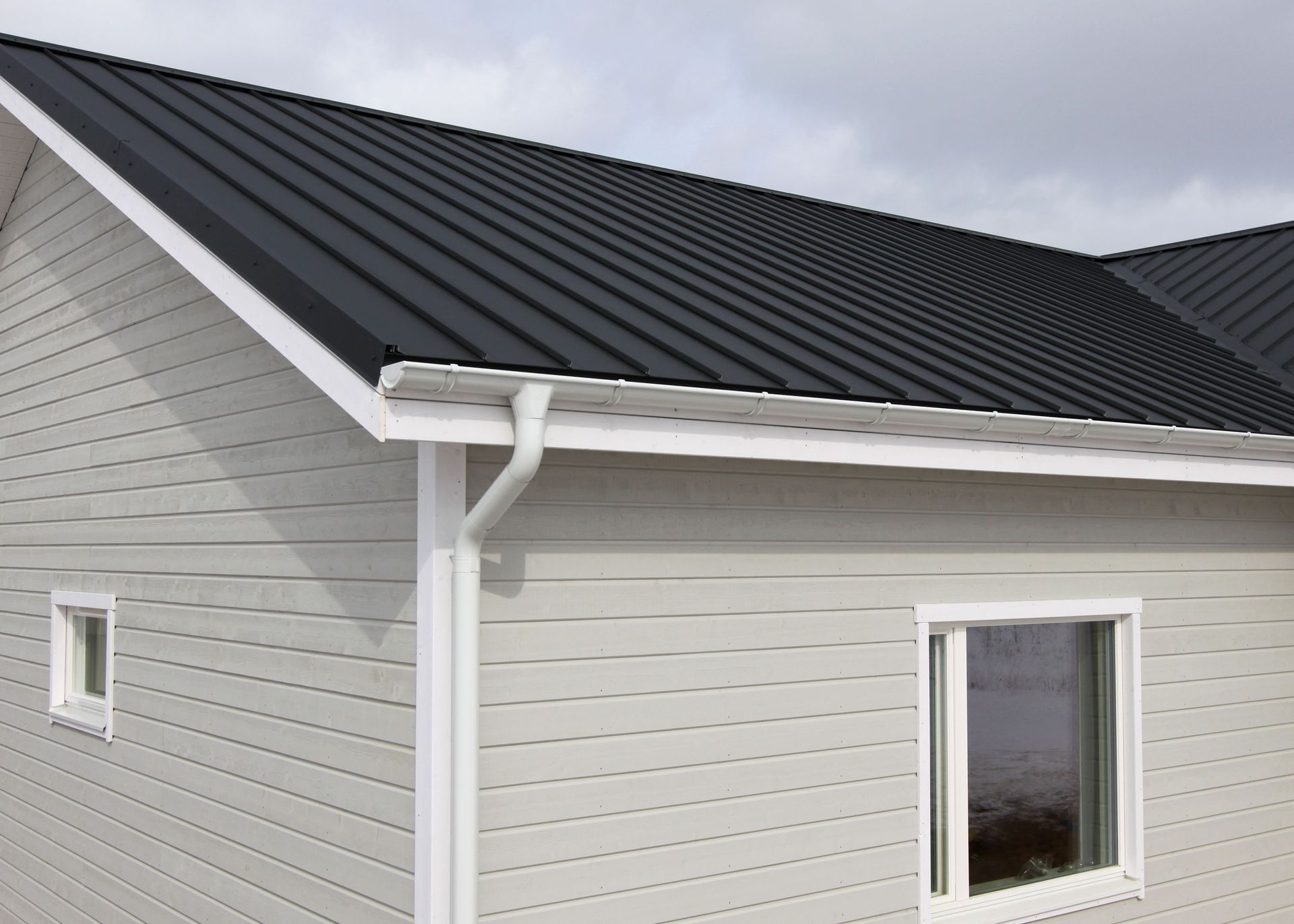 Gray house with a black metal roof, white trim, and a white gutter under an overcast sky.
