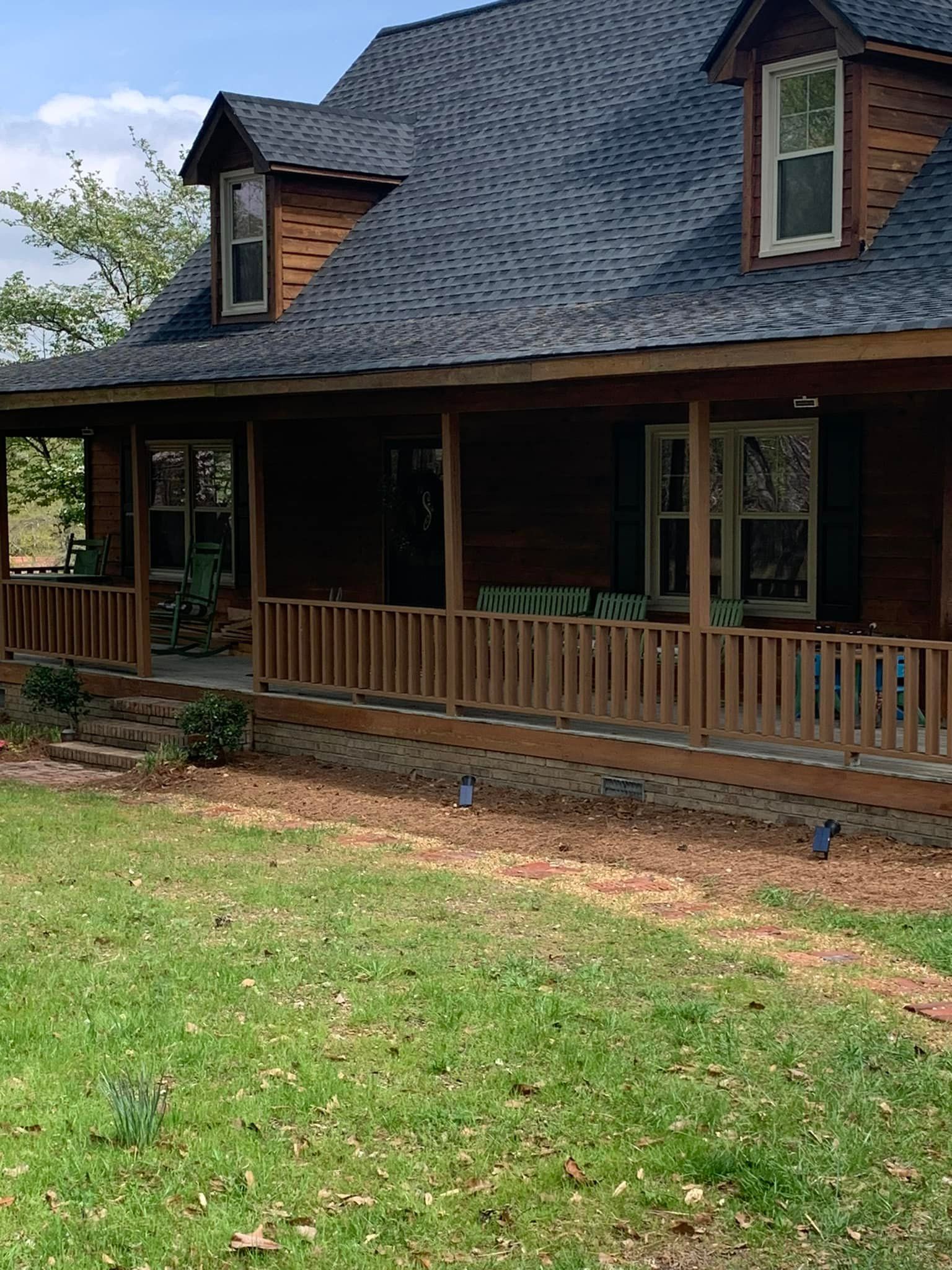 Brown house with a large porch and dormers, set against a green lawn and blue sky.