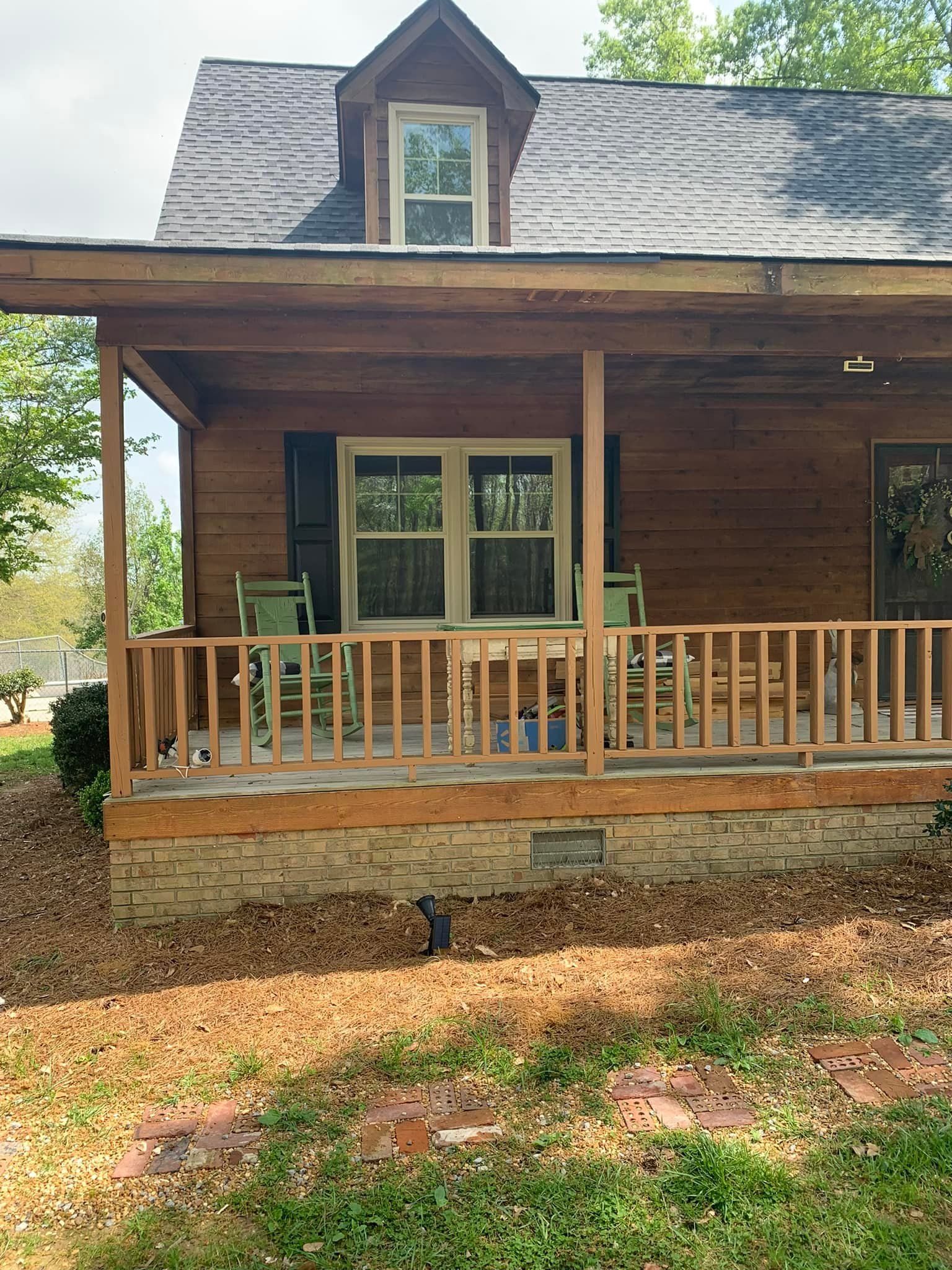 Wooden-sided house with a porch. Green chairs sit on the porch. Brown leaves and brick around the base.