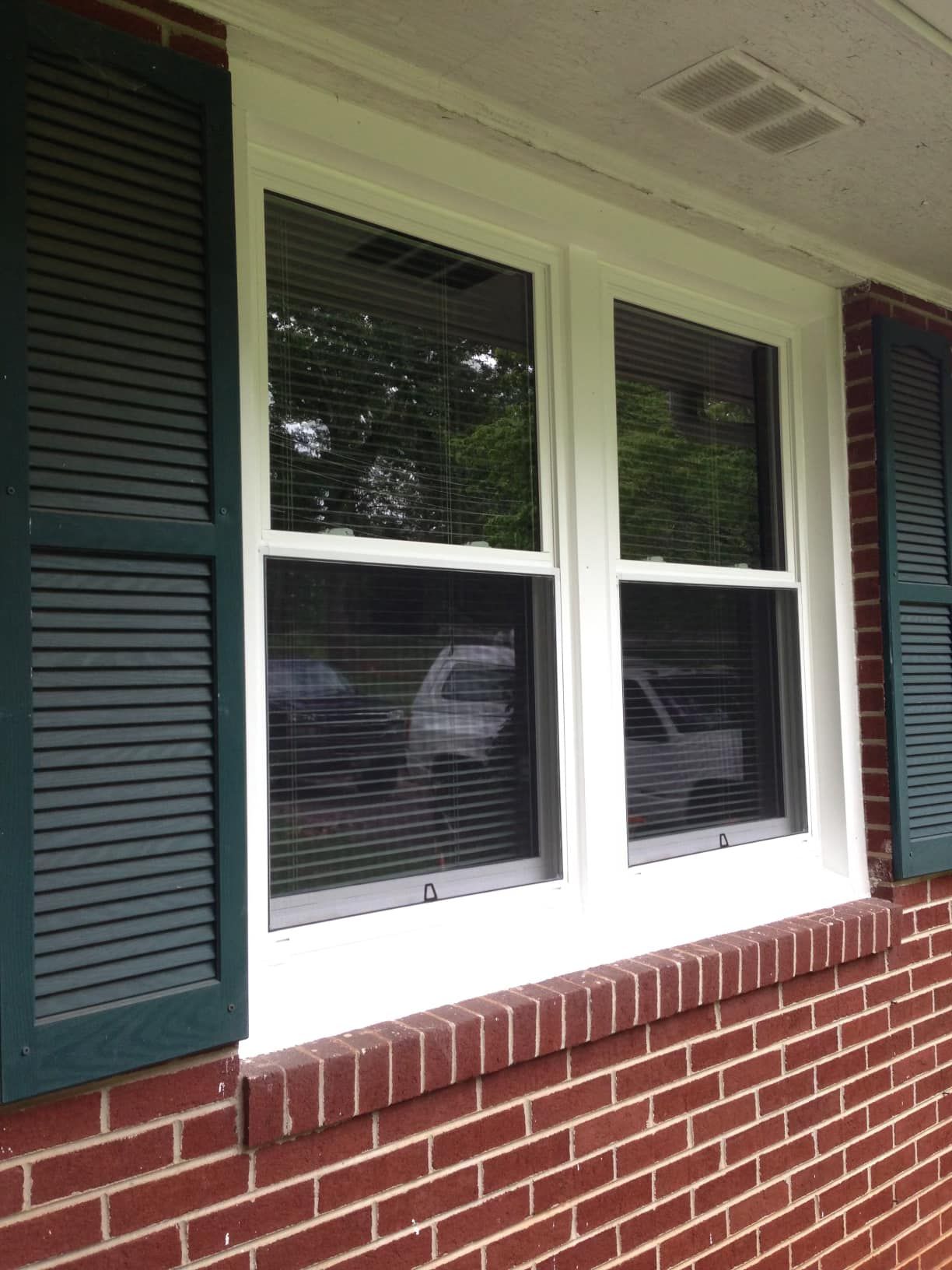 White-framed window with green shutters on a brick wall. A car is visible through the window blinds.