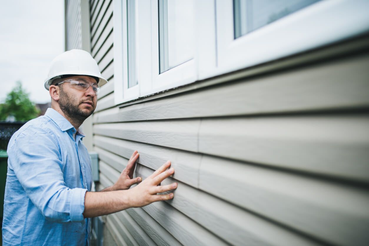 Man in hard hat examines siding on a house exterior, near a window.