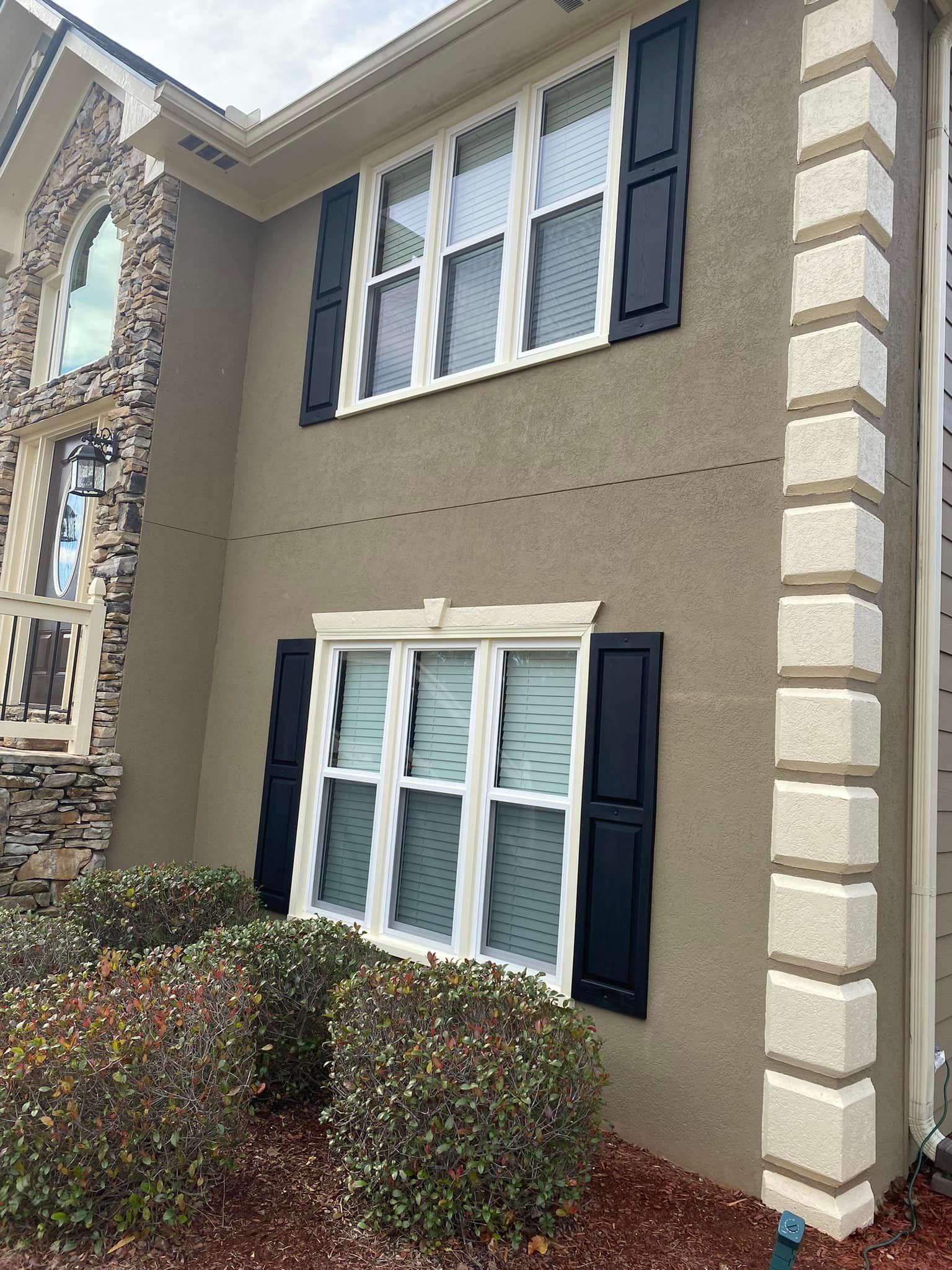 Tan stucco house with black shutters, white-framed windows, and stone detailing. Landscaping in front.
