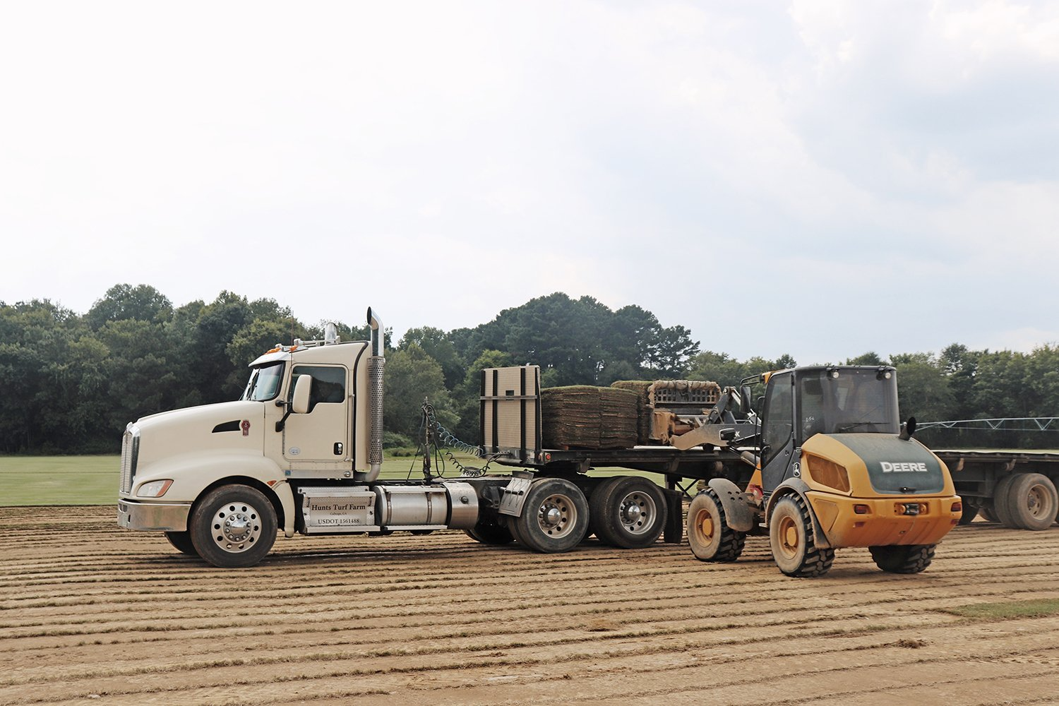 White semi-truck and yellow construction vehicle next to a trailer loaded with wooden pallets in a field.