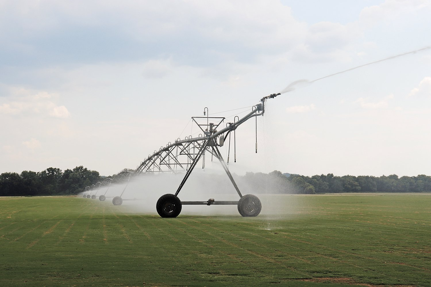 Center-pivot irrigation system spraying water over a green field, with trees in the background under a cloudy sky.