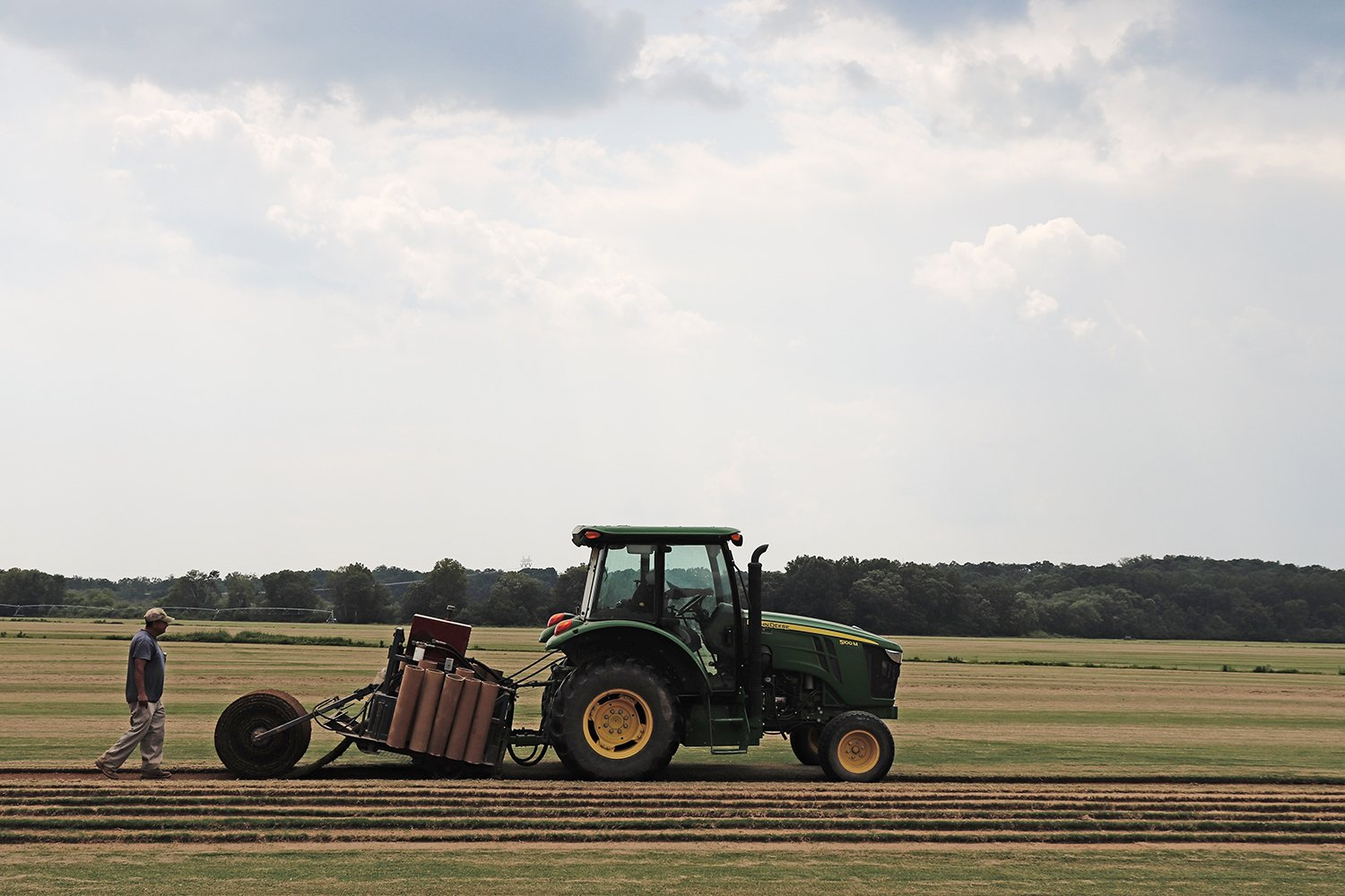 Man walking behind a green tractor, planting crops in a field under a cloudy sky.