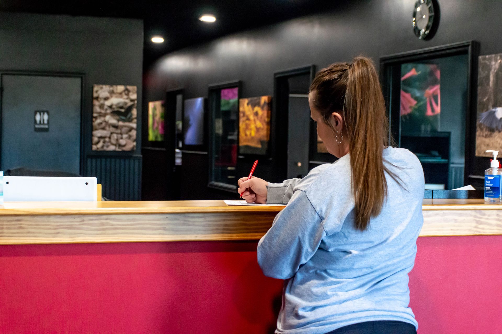 A person stands at a service counter with a red front, writing on a document in a room with dark walls and wall art.