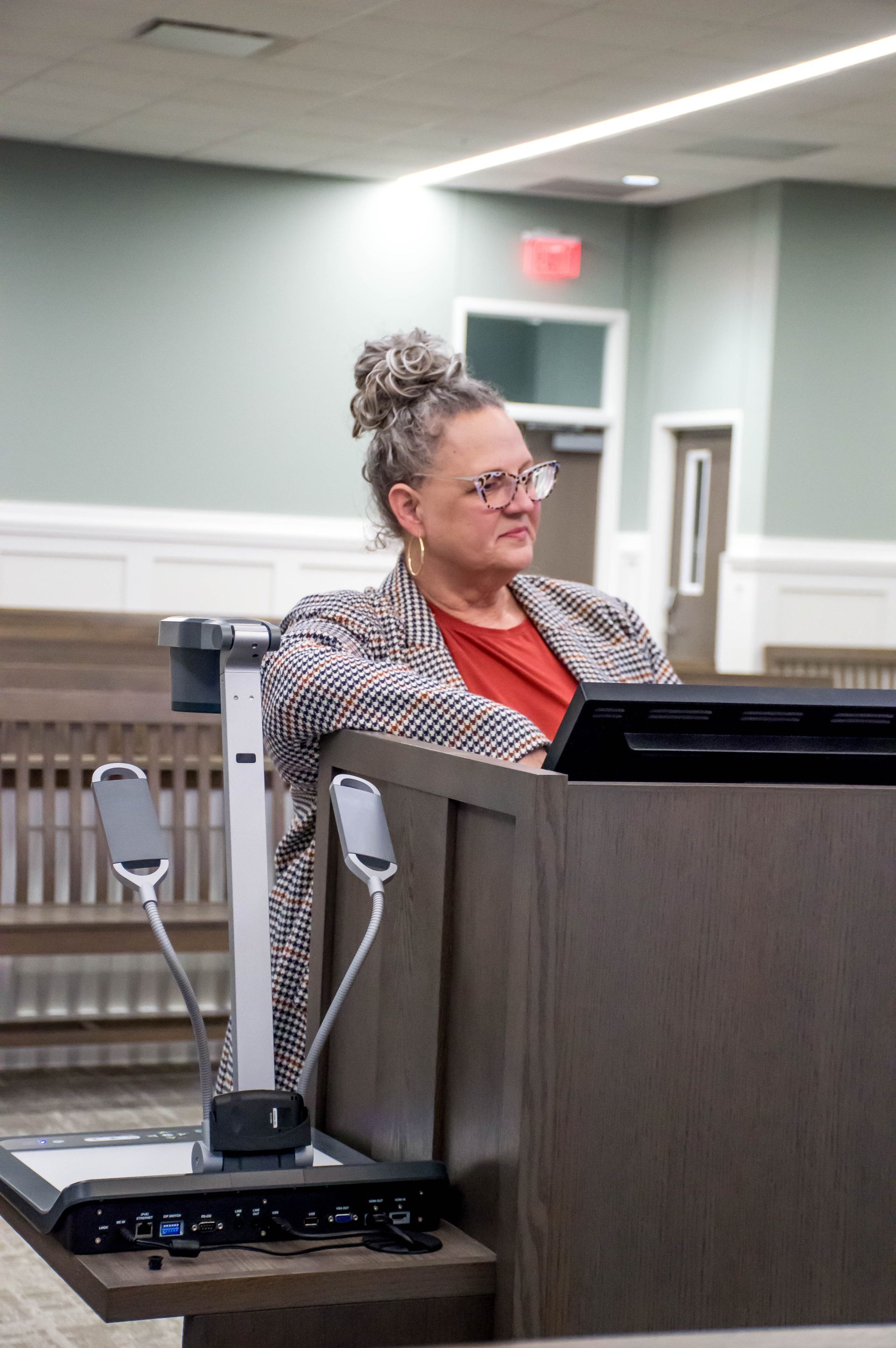 A person with glasses and hair in an updo stands behind a podium in a room with light green walls and wooden benches.
