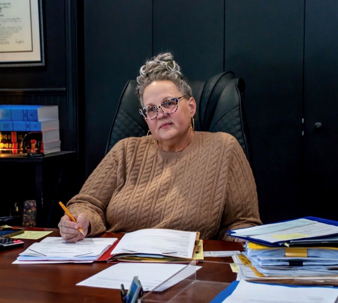 A person in a brown sweater sits at a desk, holding a pen and looking forward with documents spread across the workspace.