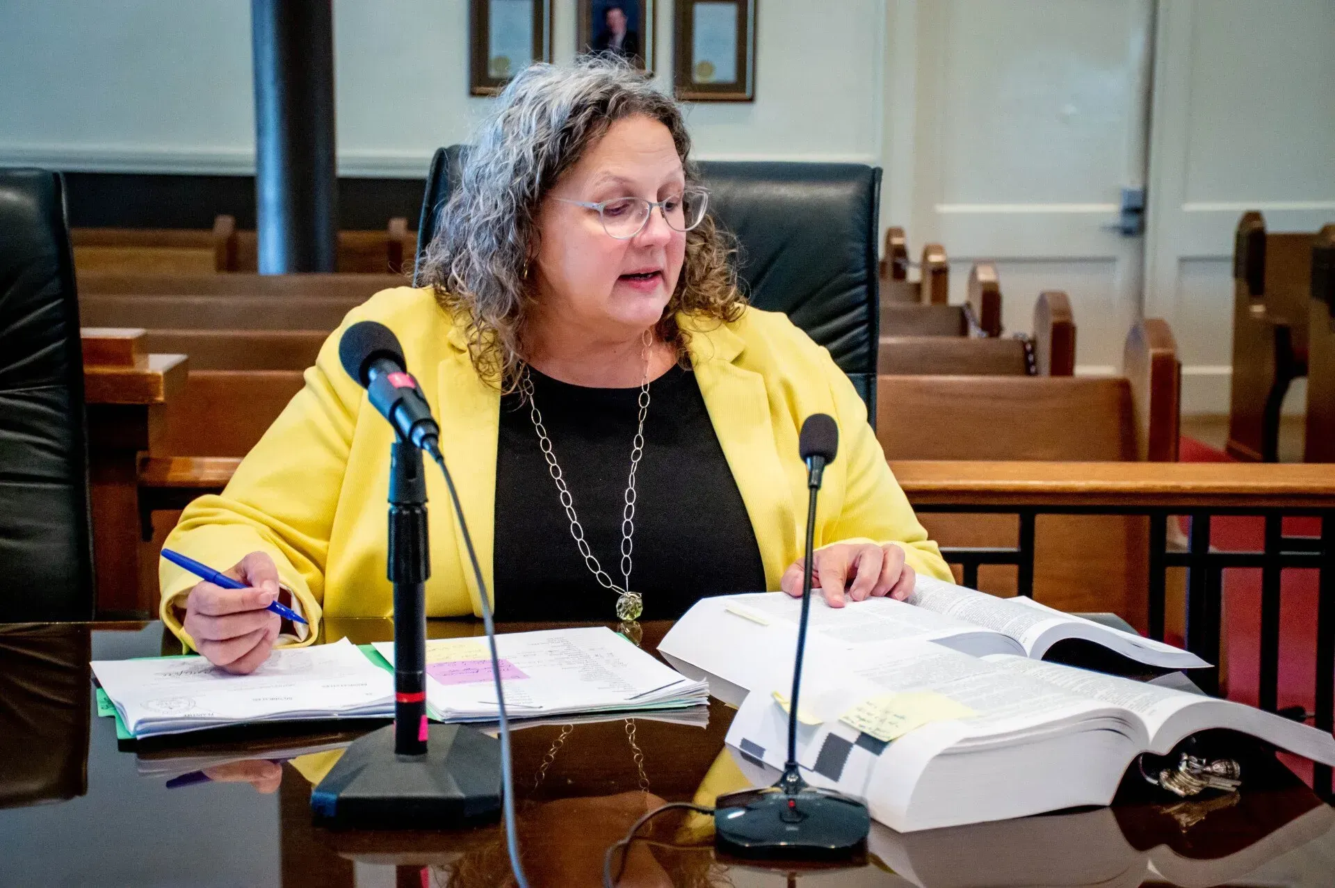 Woman in yellow blazer at a desk, reviewing documents, microphones present.