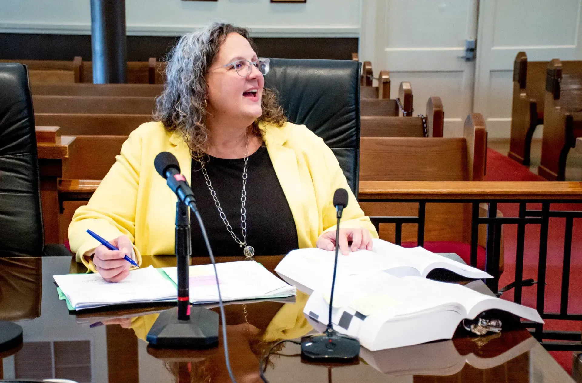 Woman in a yellow blazer speaks at a desk with microphones, documents, and books in a courtroom setting.