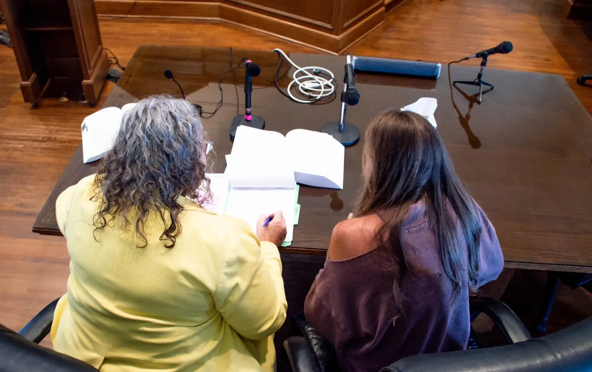 Two people seated at a table with papers, microphones. Interior. One person writes.