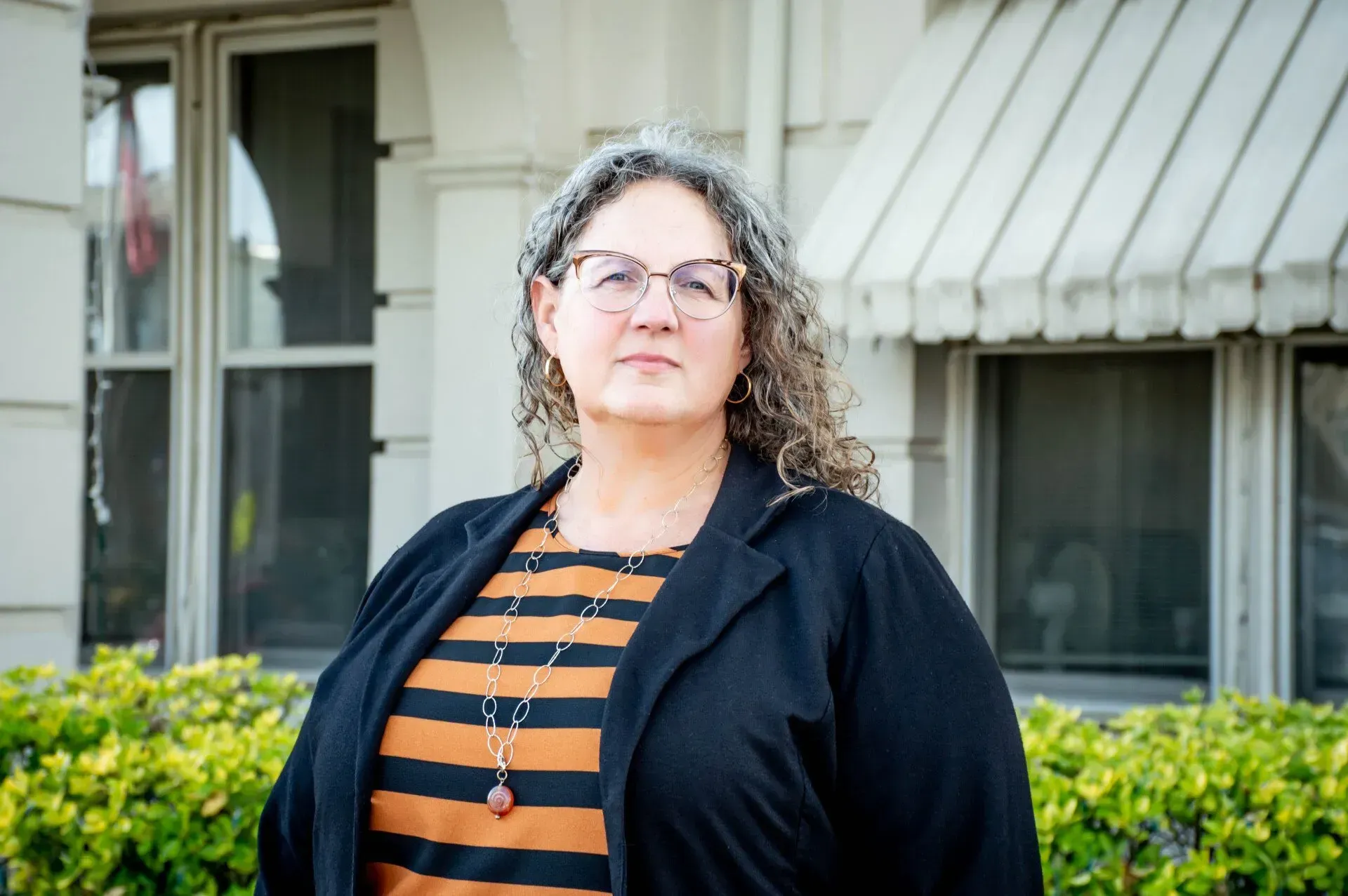 Woman with glasses and gray curly hair stands in front of a white building, wearing a striped shirt and black jacket.