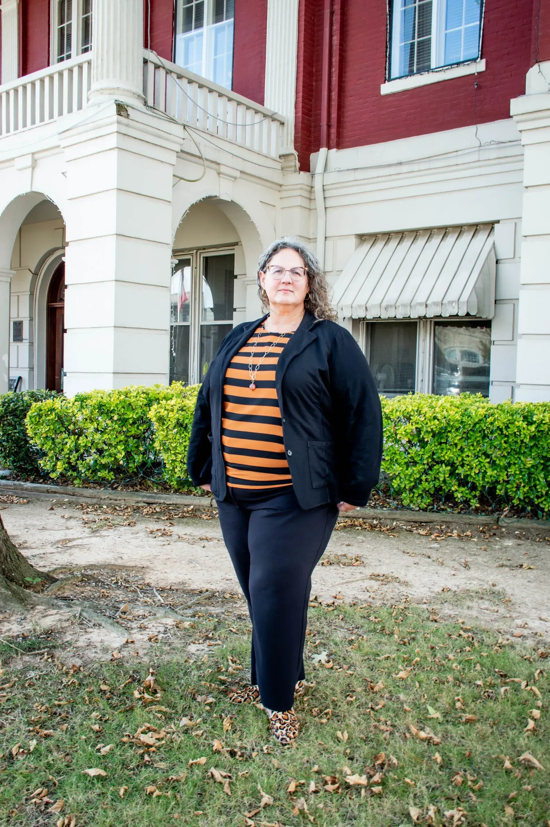 Woman in black blazer and pants, standing in front of a red building with white pillars and green bushes.
