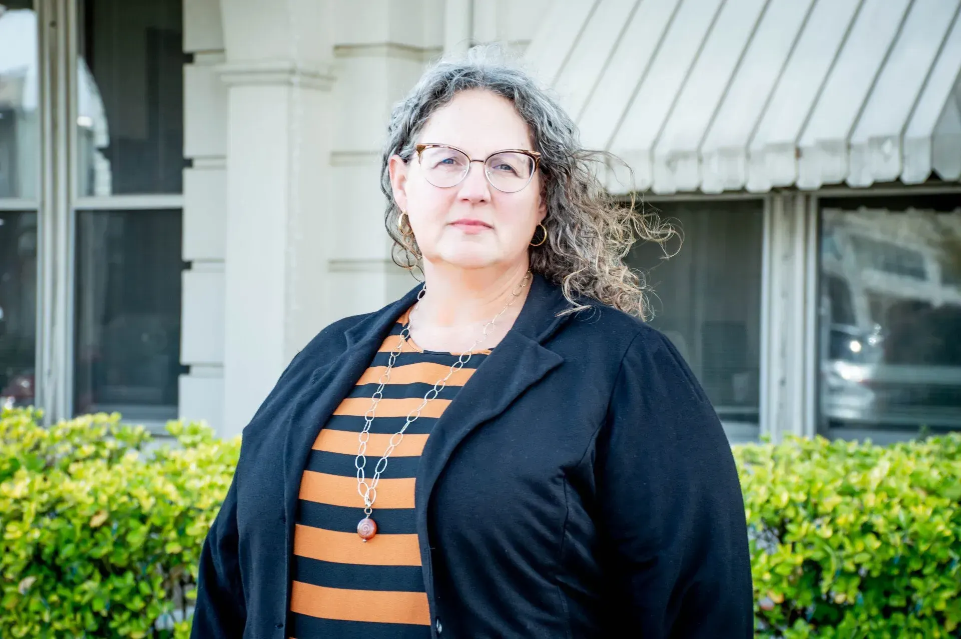 Woman with glasses and graying hair, wearing black jacket and striped shirt, standing outside a building.