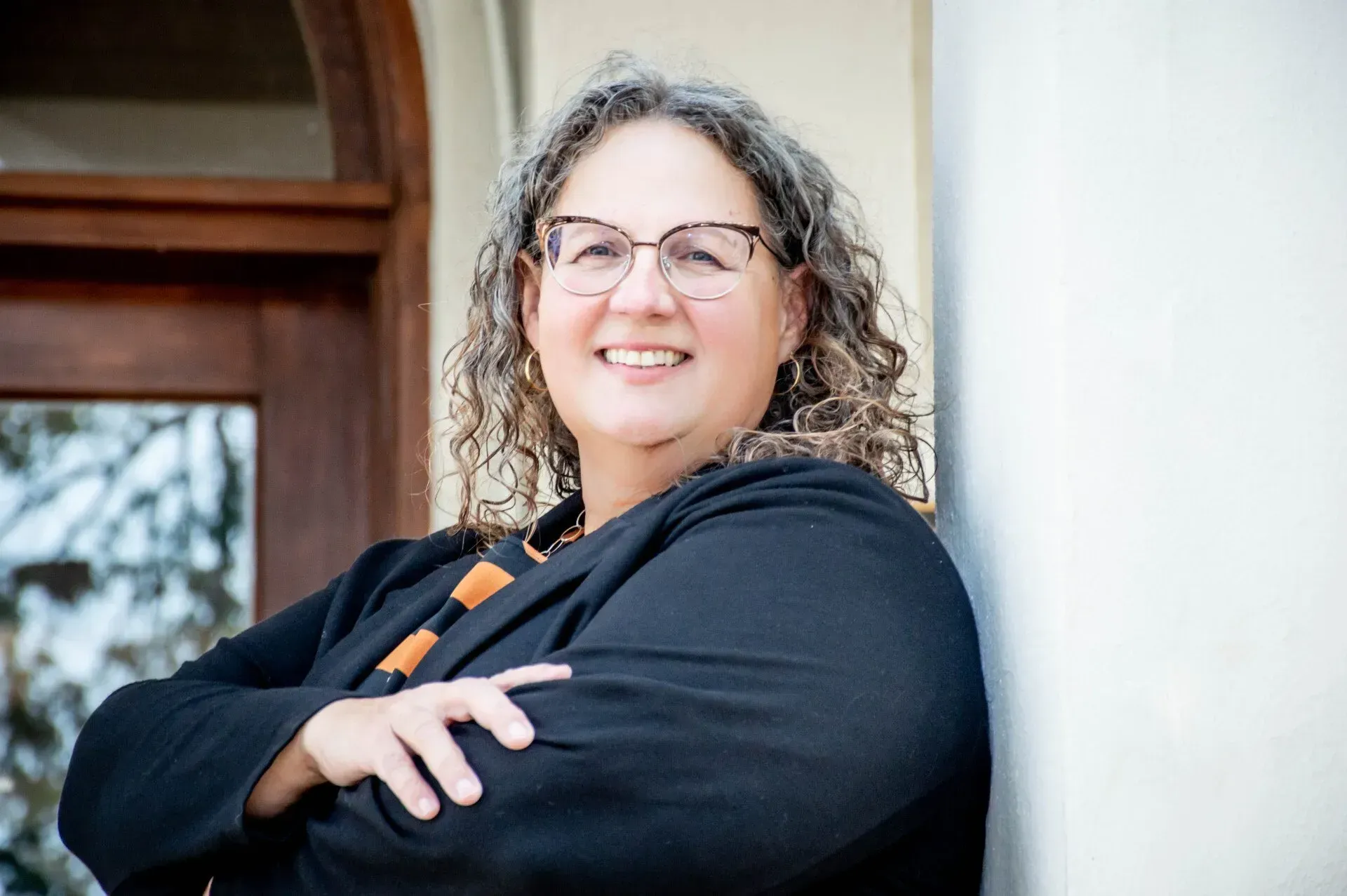Woman with glasses, smiles, arms crossed, leaning against a white pillar, black blazer, outdoors.