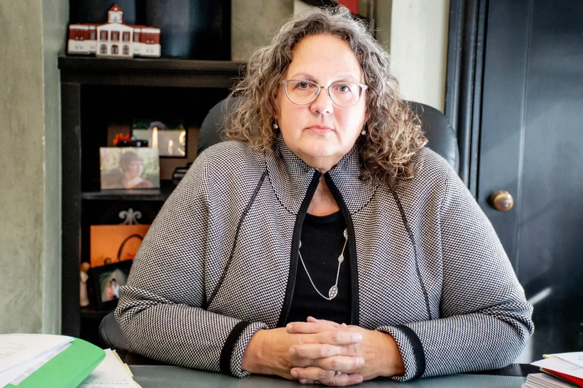 Woman in glasses, patterned jacket, sitting at a desk in an office setting.