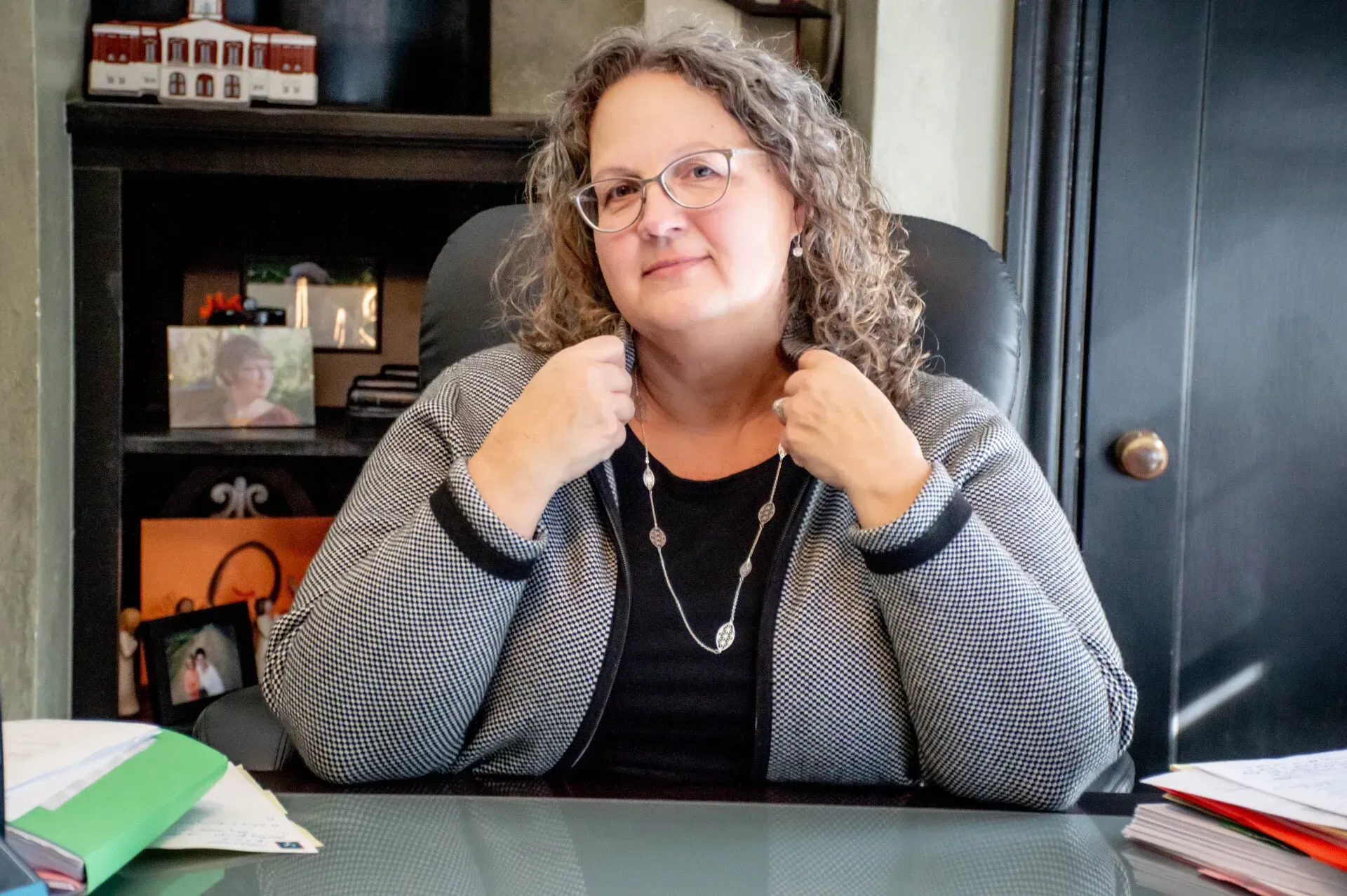 Woman in glasses, black shirt, and patterned jacket sits at desk. Background includes shelves, photos, and a door.