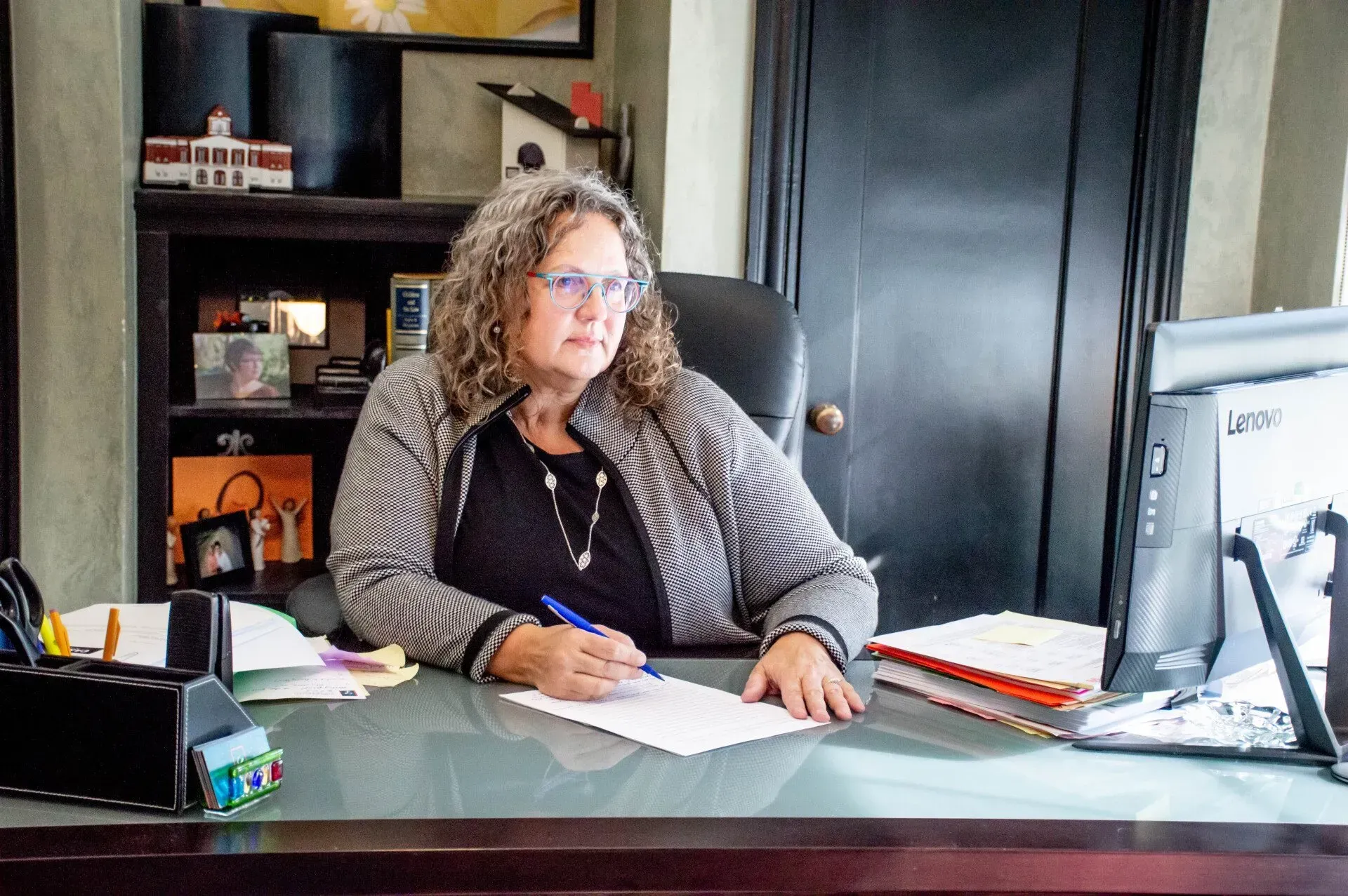 Woman in a blazer sits at a desk, writing on paper in an office.