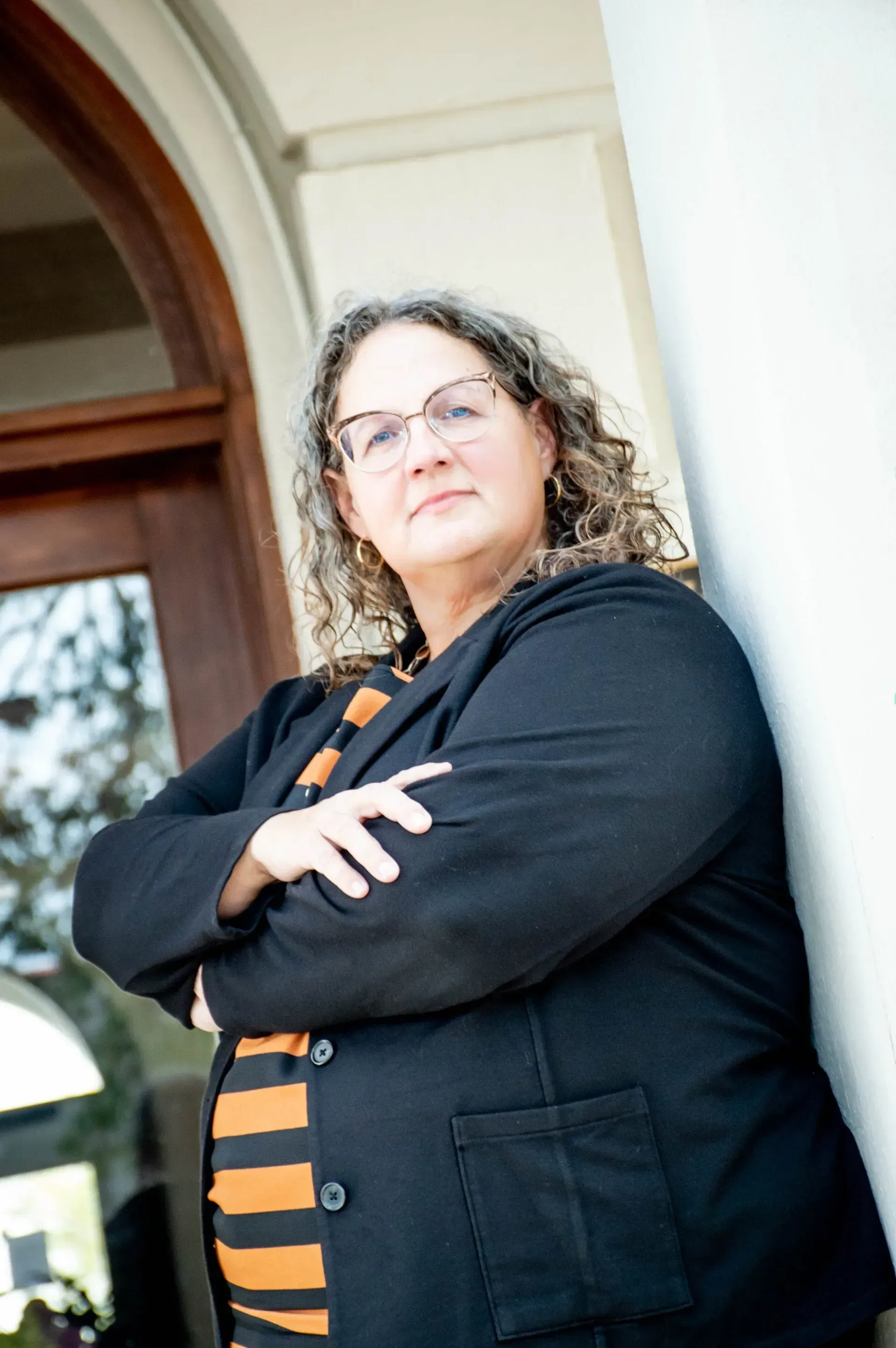 Woman with glasses and curly hair, arms crossed, leaning against a white column near a wooden door.