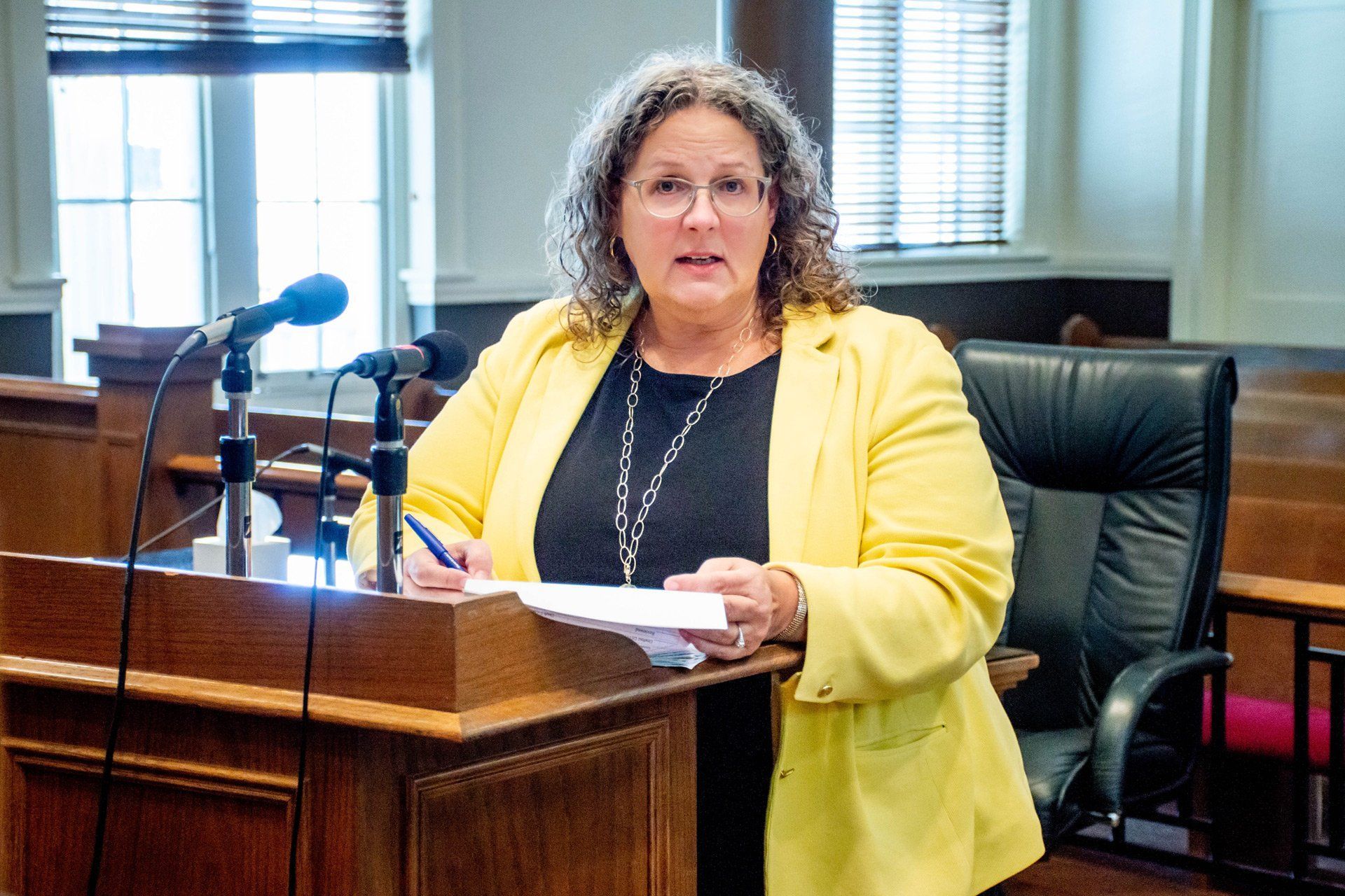 Woman in yellow jacket speaks at a wooden podium in a courtroom.