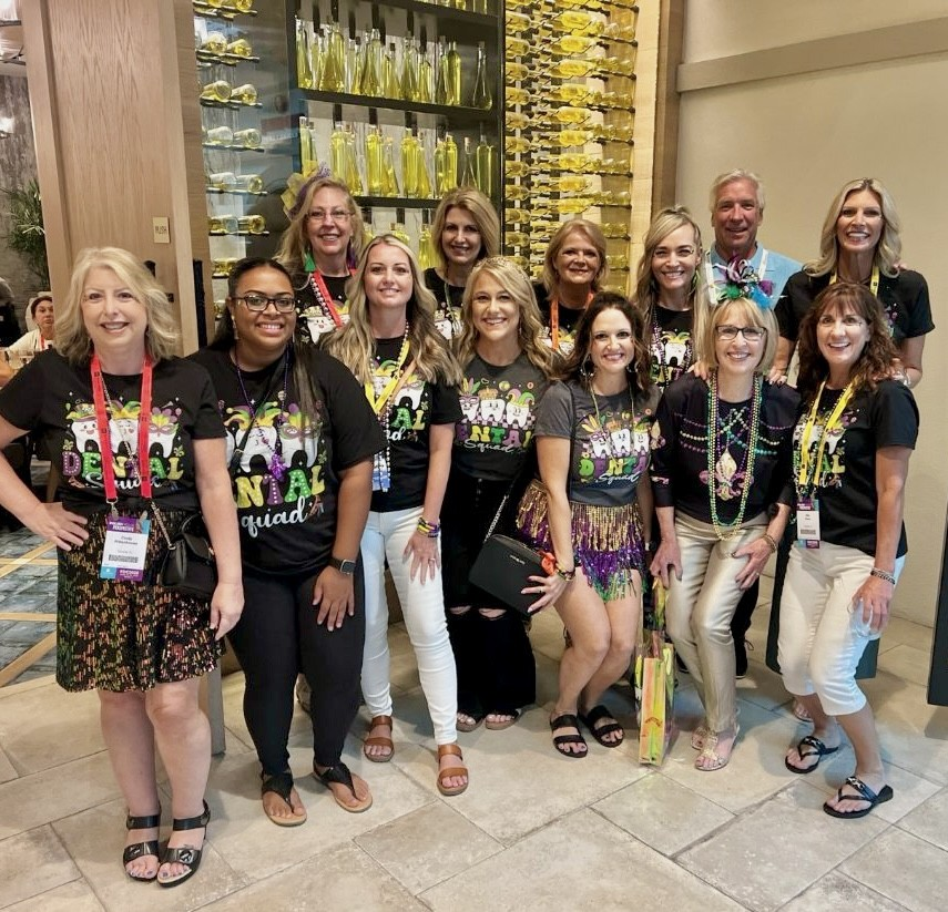 Group of smiling people posing indoors in matching black shirts and leis, with shelves of yellow bottles behind them