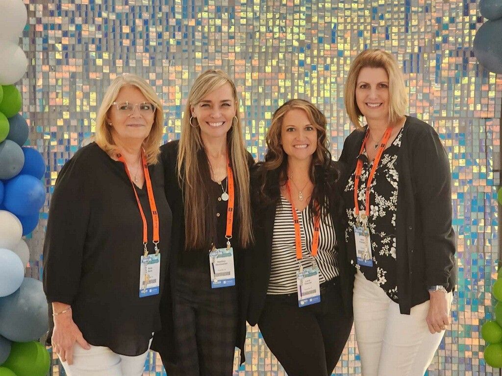 Four people smiling at an event, standing before a colorful mosaic wall and blue balloons.