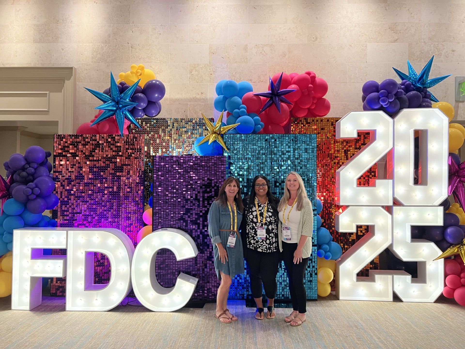 Four people posing beside large “FDC” and “2025” letters with colorful balloons and a glittery backdrop