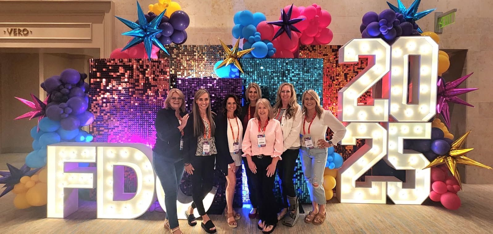 Group posing at a colorful event backdrop with large “FD” and “2025” letters and decorative flowers