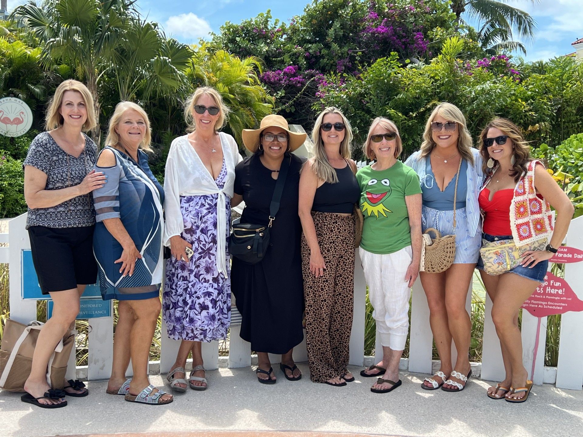 Seven women posing outdoors in front of lush greenery and colorful flowers, smiling in summer outfits