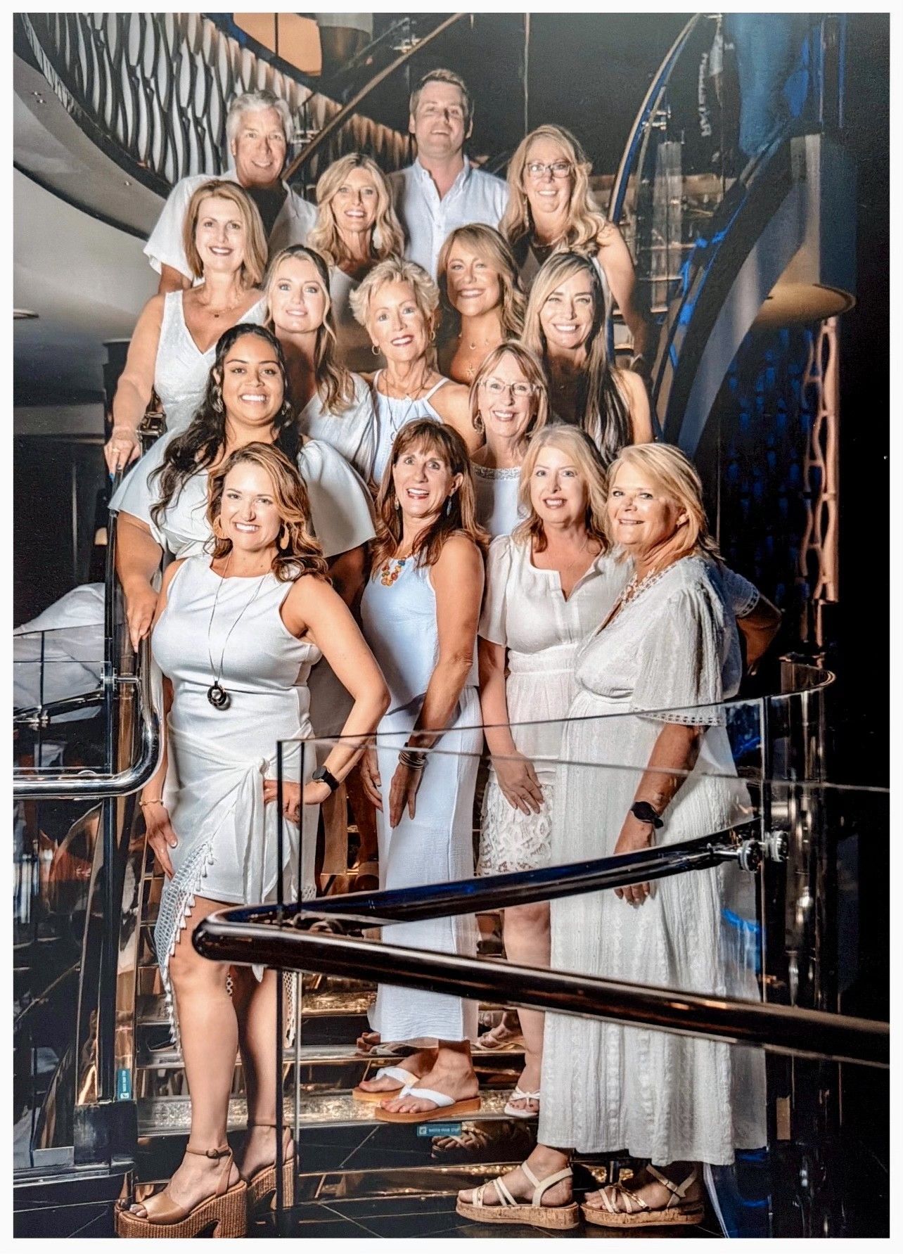 Group of people posing on a staircase in elegant white outfits, smiling in a formal indoor setting