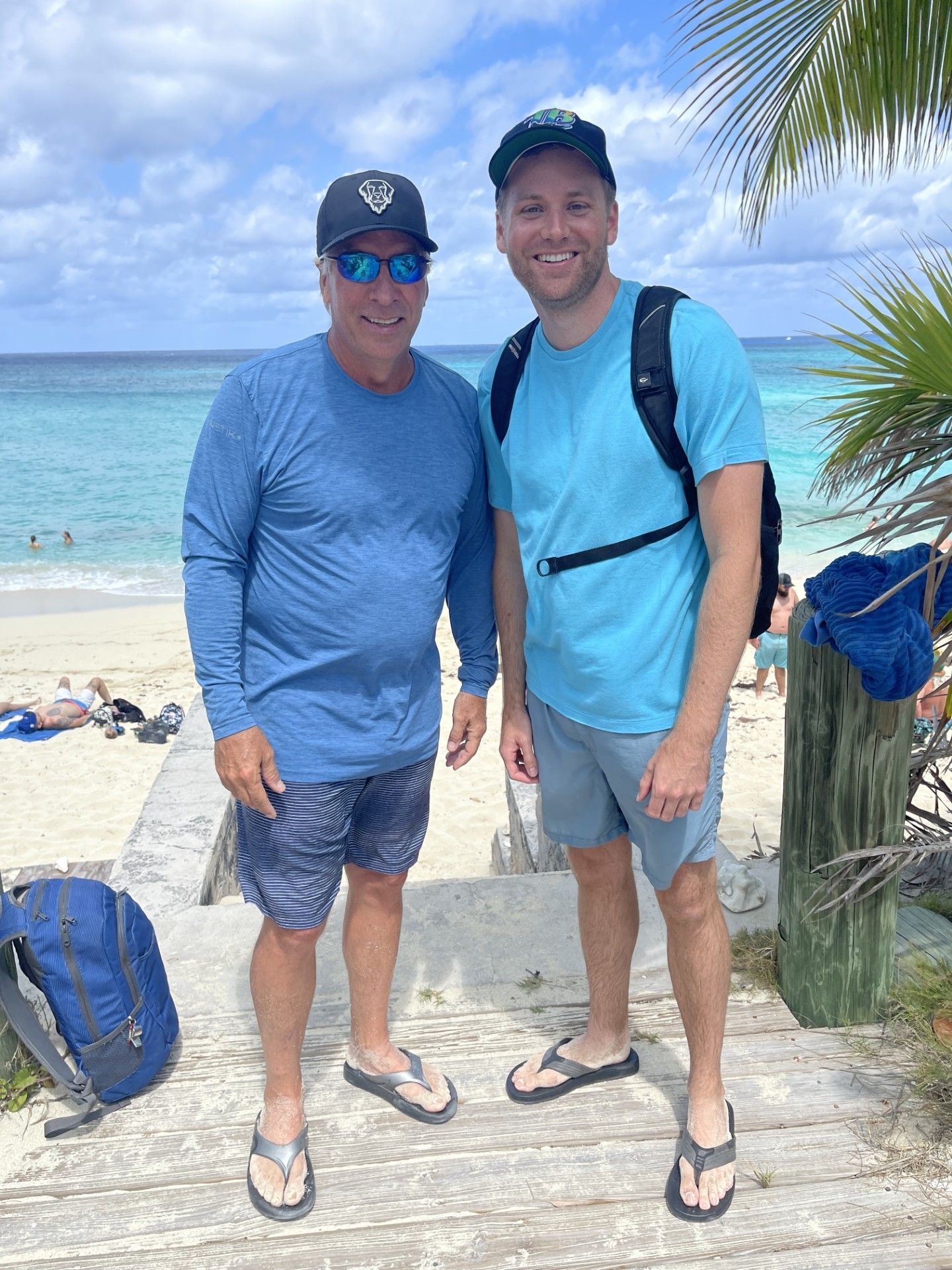 Two people standing on a beach boardwalk with turquoise water and palm leaves in the background.