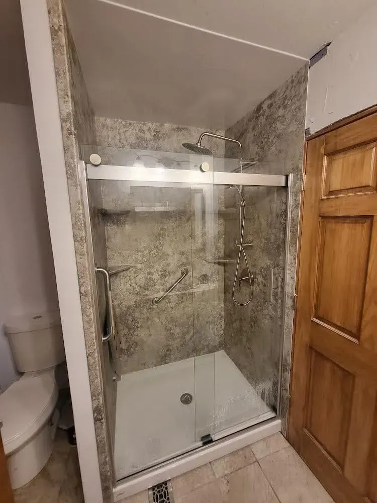 Shower stall with stone-like walls, glass doors, and multiple showerheads; next to a toilet and wooden door.