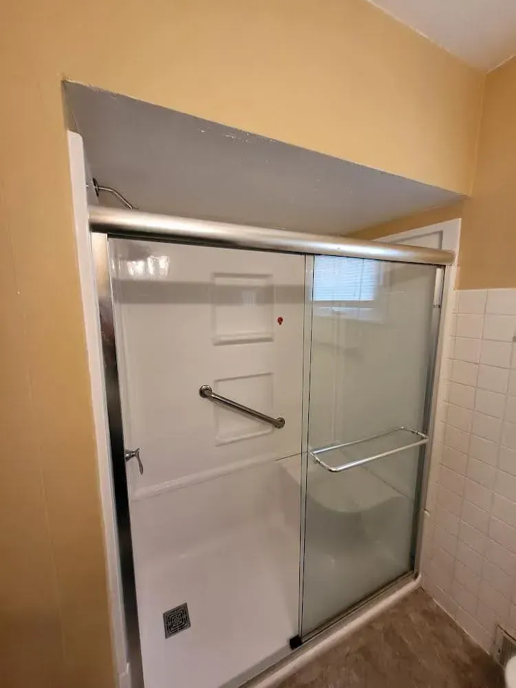 White shower stall with glass sliding doors and a chrome handrail, set in a beige-colored bathroom.