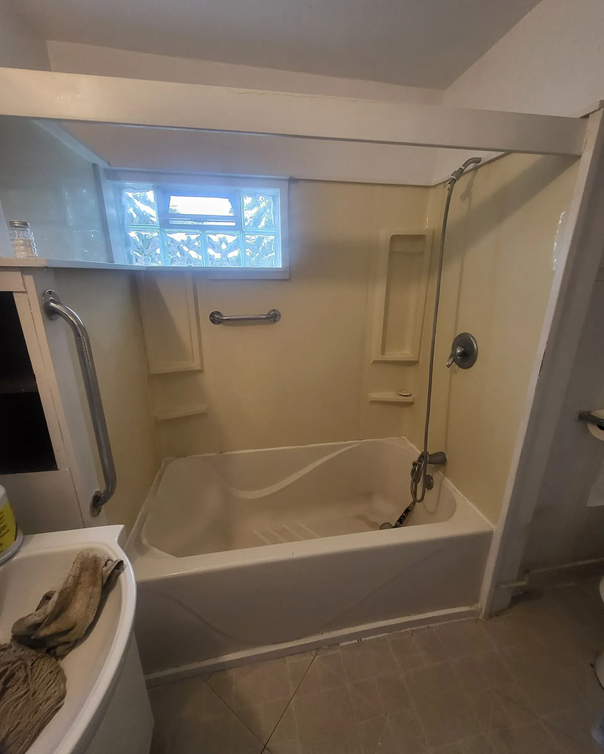 Bathroom with bathtub, grab bars, and showerhead; beige walls and light from a small window.