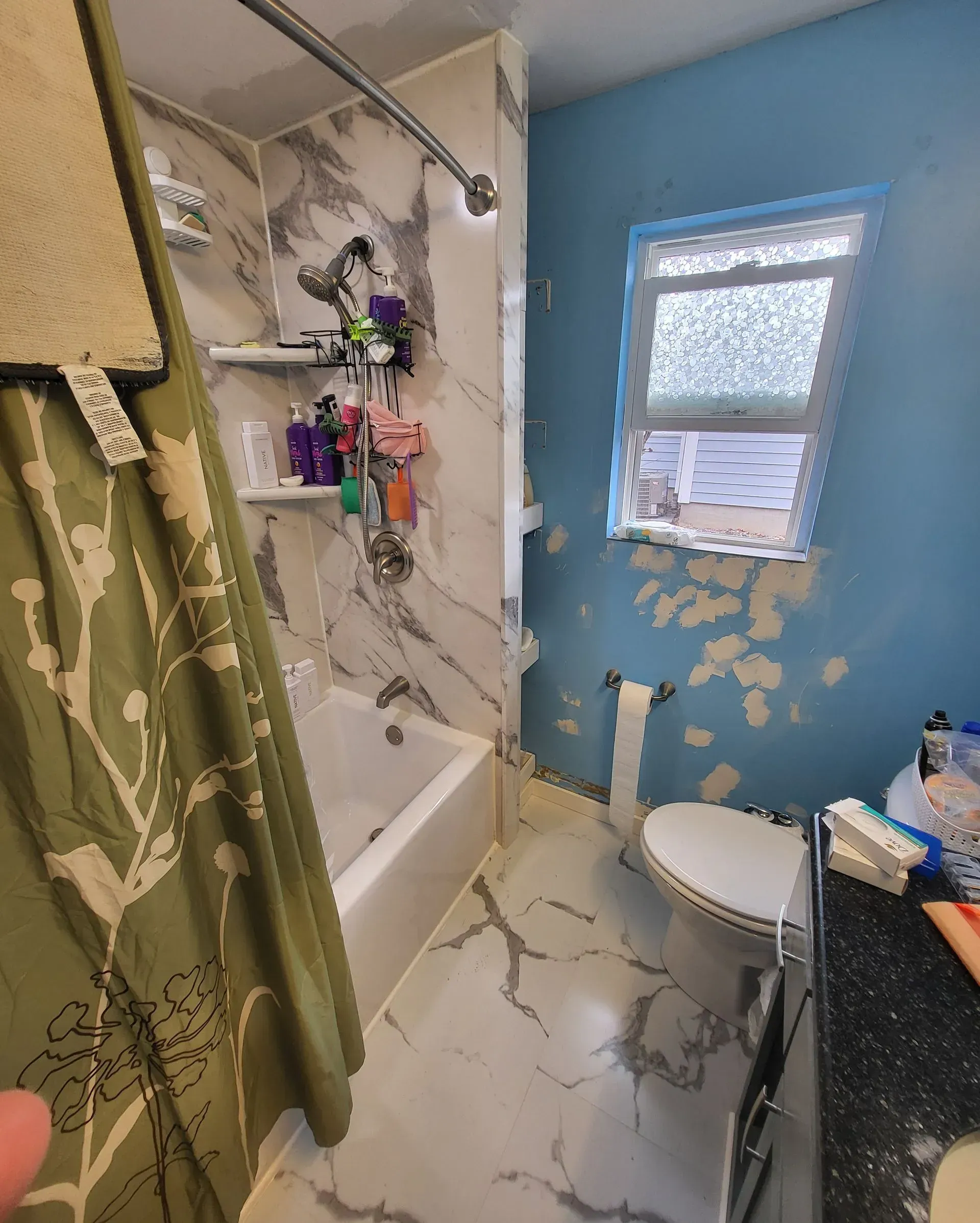Bathroom with marble-look shower, white tub, toilet, and blue wall with peeling paint.