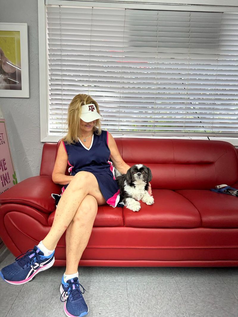 Woman and small dog on a red couch near a window; the woman is wearing a hat and running shoes.
