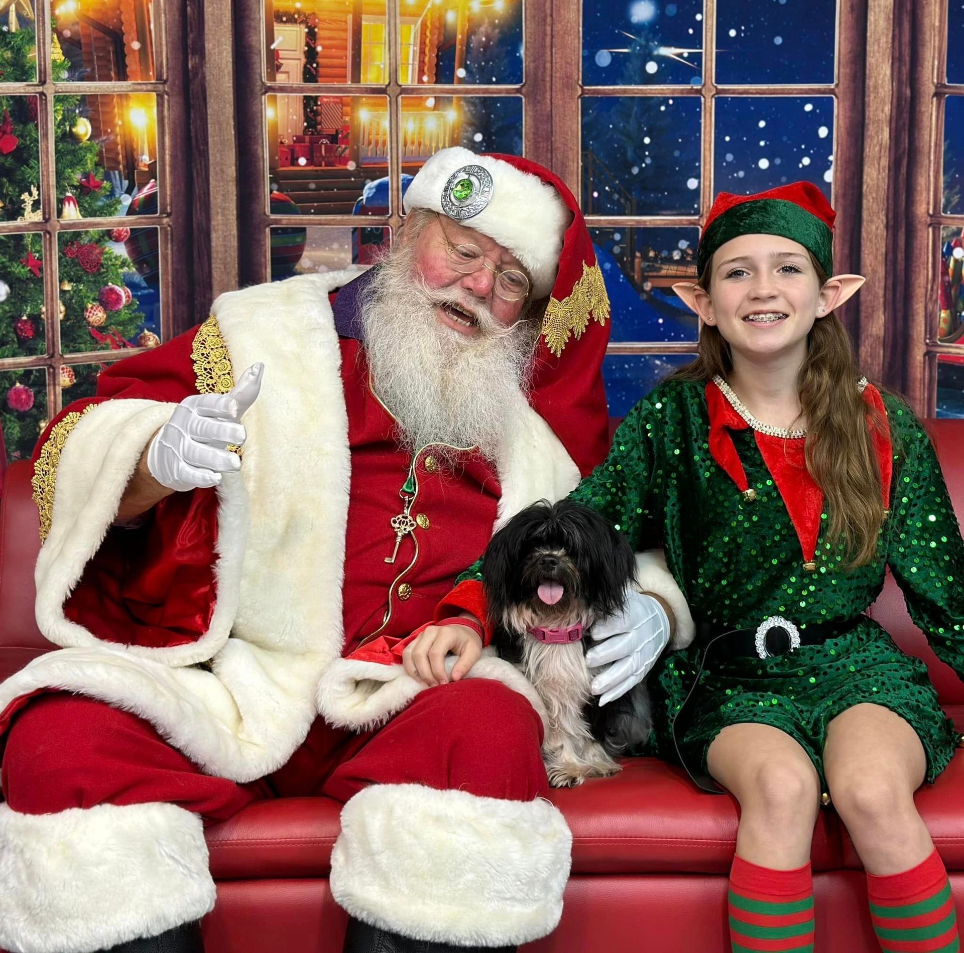 Santa Claus, a young elf, and a small dog sitting on a red couch. Santa points upward, smiling.