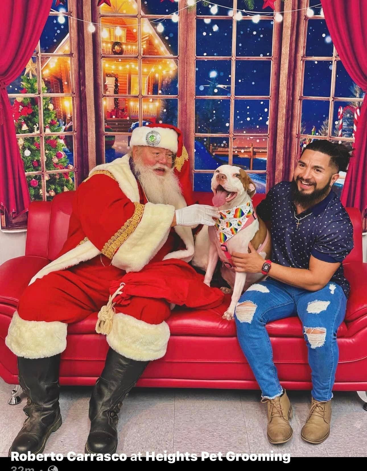 Santa, a man, and a dog pose on a red couch. Holiday backdrop. The dog smiles.